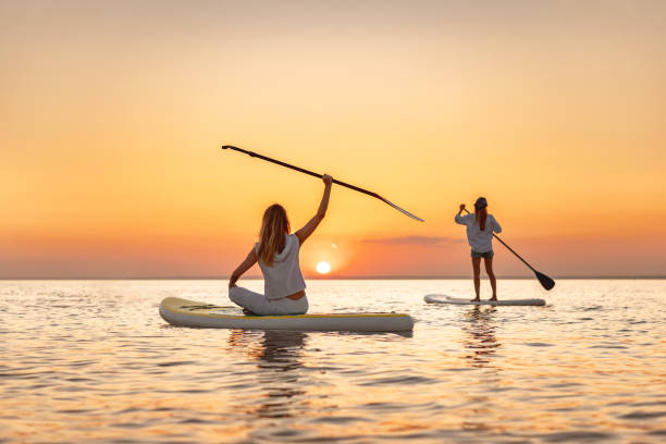 Two happy slim young girls walks at sunset lake on sup stand up paddle boards